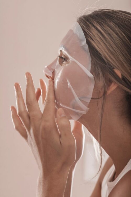 Frau legt eine weiße Tuchmaske auf ihr Gesicht, Seitenansicht – Anwendung einer Exosome Sheet Mask für intensive Hautpflege.
